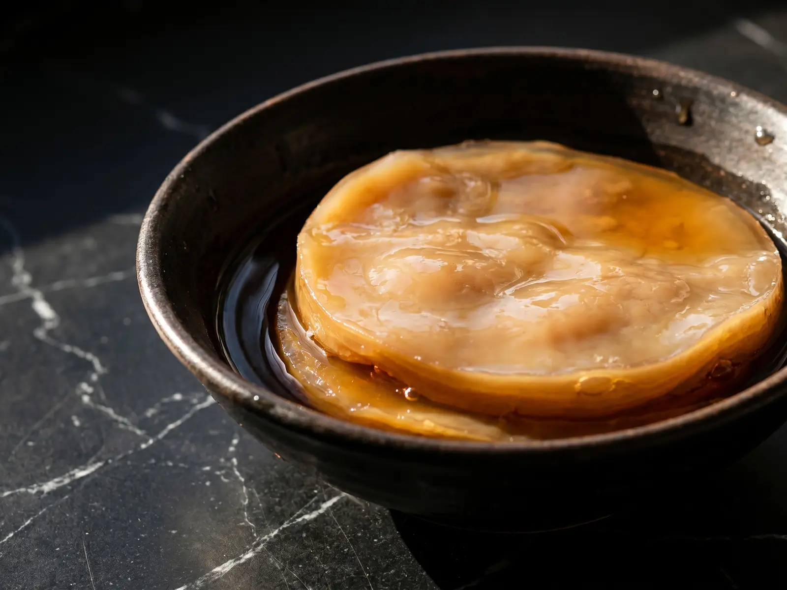 kombucha valeur nutritionnelle Bol de thé sombre avec un disque de SCOBY beige-brun sur marbre noir, éclairé par une lumière naturelle.