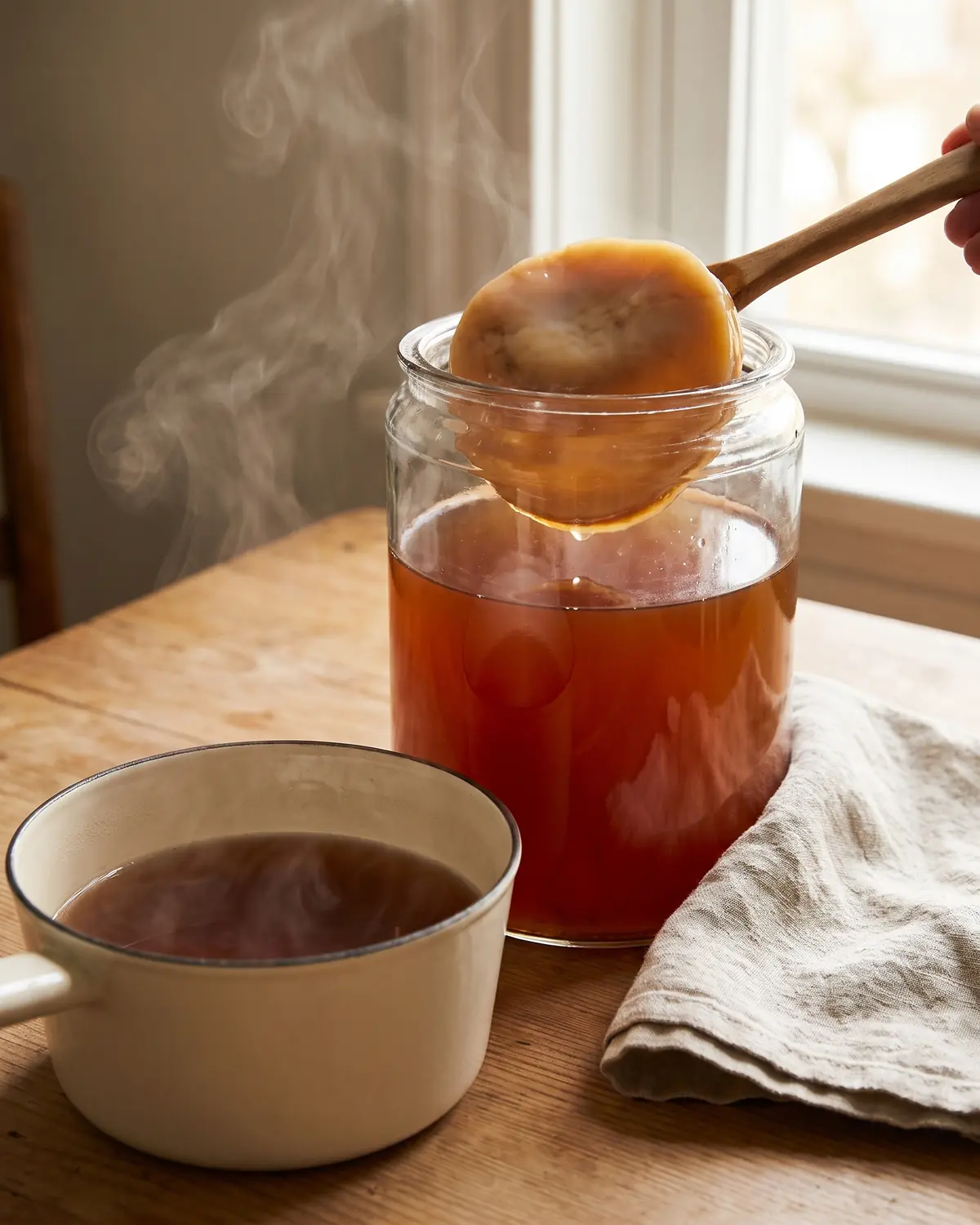 Un filet de vapeur s&rsquo;&eacute;l&egrave;ve d&rsquo;une casserole &eacute;maill&eacute;e de th&eacute;, pr&egrave;s d&rsquo;un bocal de SCOBY pos&eacute; sur du lin.