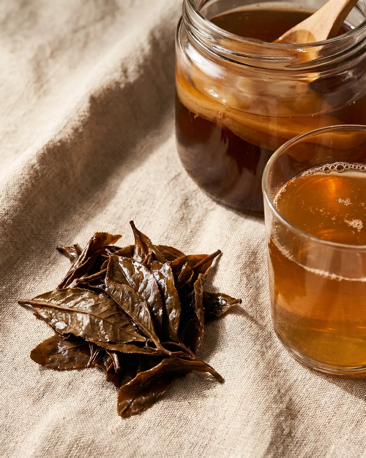 Feuilles de th&eacute; humides pr&egrave;s d&rsquo;un verre de kombucha sur une surface en lin naturel.