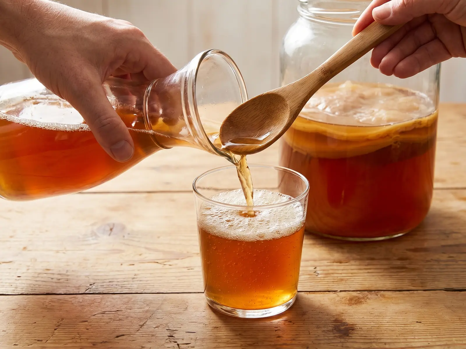 Main servant du kombucha ambr&eacute; dans un verre, avec une fine mousse sur une table en bois clair.
