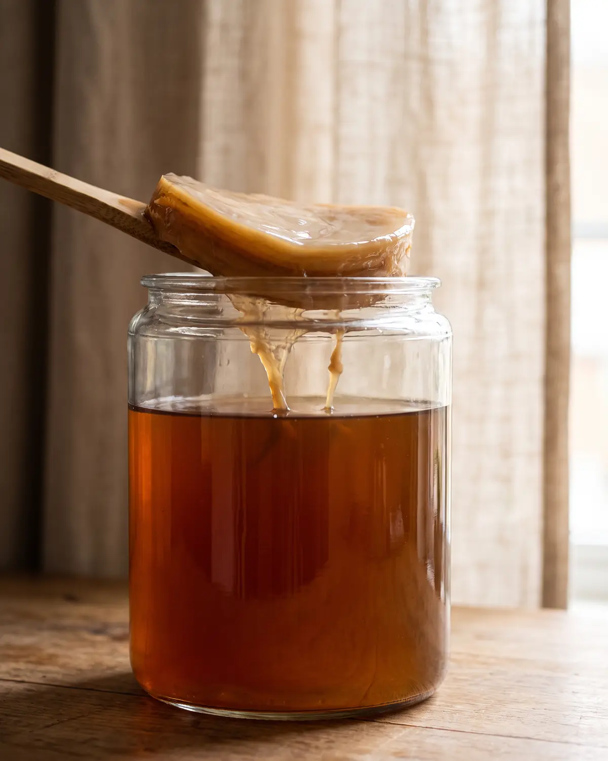 difference entre kefir et kombucha Bocal de kombucha avec SCOBY et filaments doux, photographié près d’un voile de lin flou.