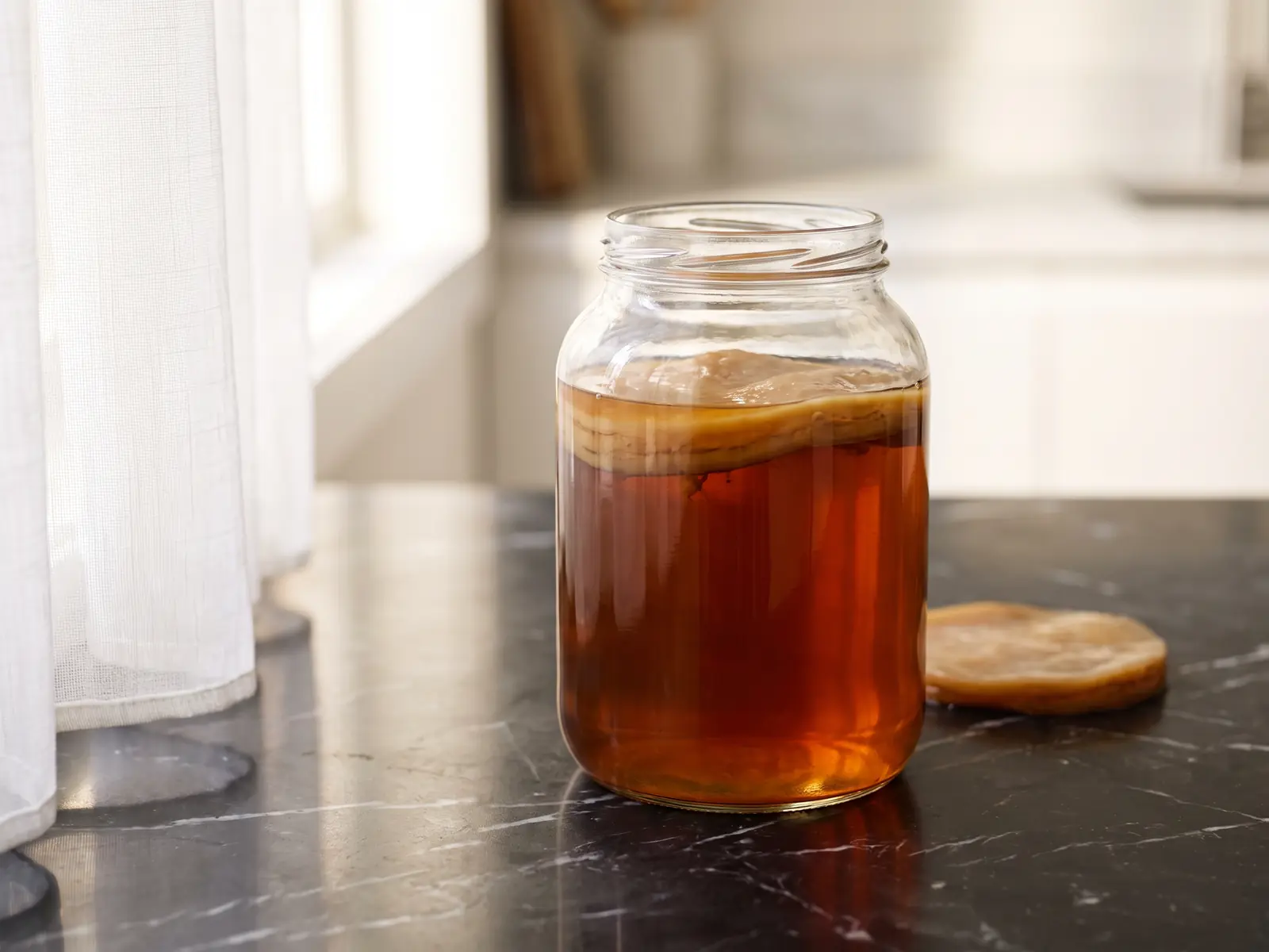 Grand bocal de kombucha ambr&eacute; avec SCOBY suspendu sur marbre noir et reflet de rideau blanc