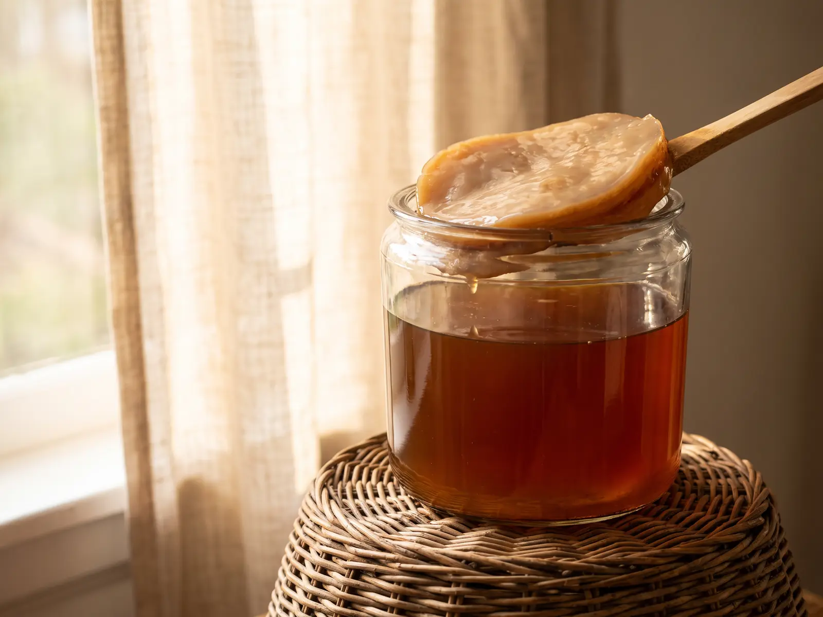 Bocal de SCOBY pos&eacute; sur un panier en osier, devant un linge clair baign&eacute; de lumi&egrave;re naturelle.